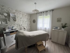 Pièce dédiée aux soins pratiqués par Elise Reflexologie. Au milieu on retrouve le lit de soin, à gauche de l'image dans le coin de la pièce un meuble avec tous les produits dédiés au soin. Toute la pièce est dans un style de décoration très doux et apaisant.